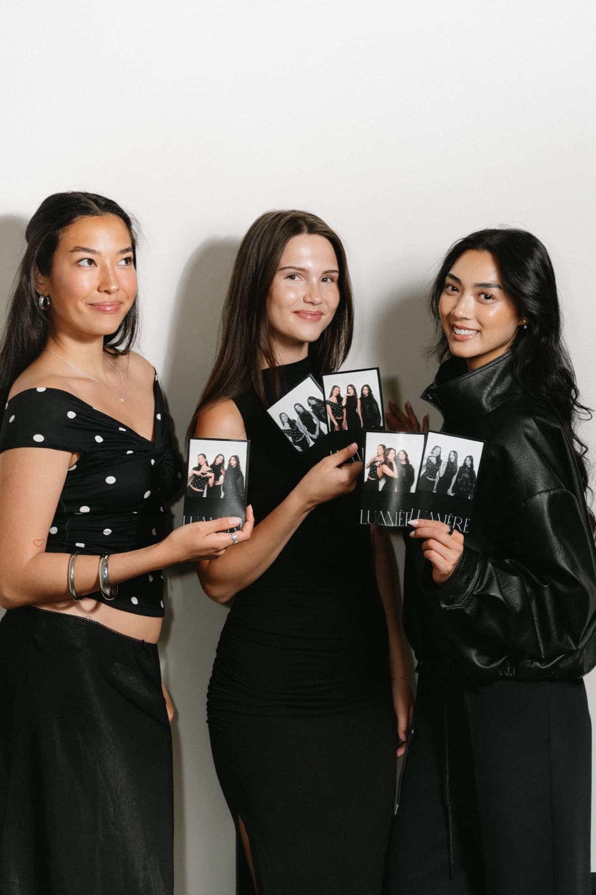 Three women in black outfits holding their black-and-white MEMORI photo strips in front of a pale studio wall.