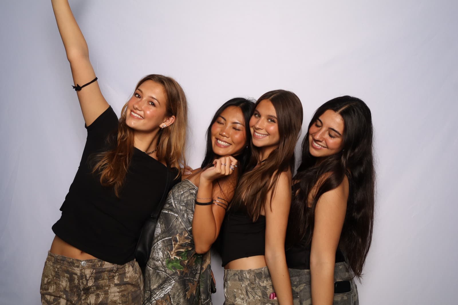 Four friends in black tops and camo posing together, one with her arm raised, in front of a pale studio wall.