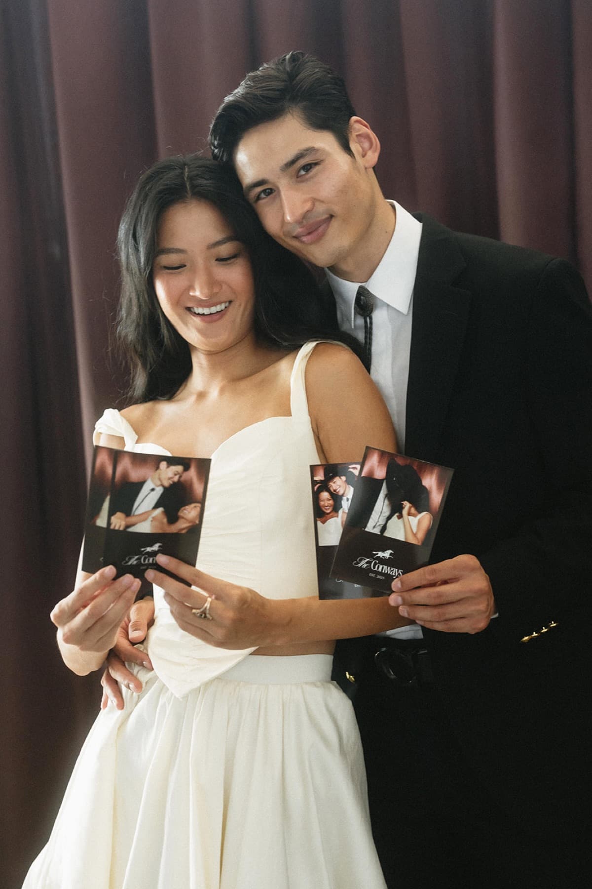A bride and groom in black tie holding their photo strips close in front of a deep burgundy curtain.