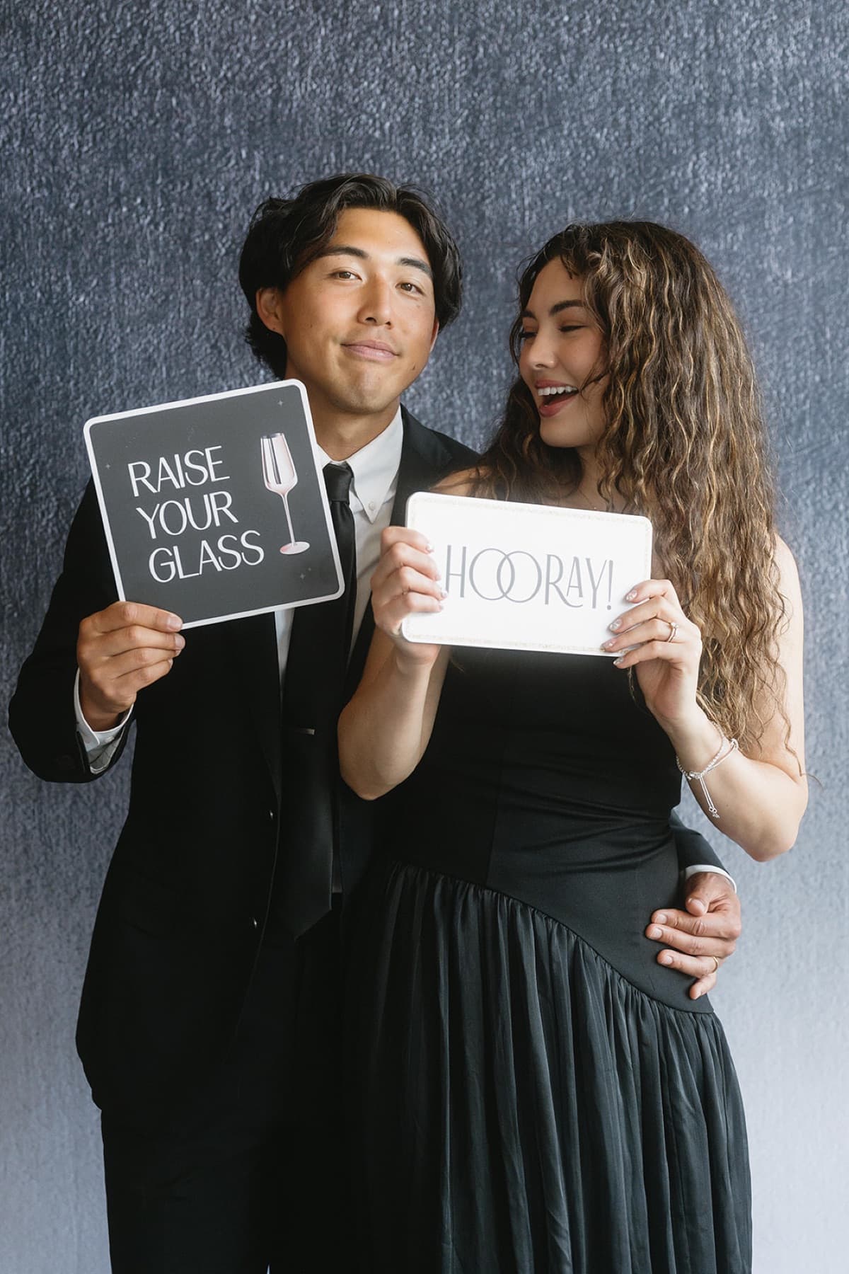 A couple in black tie holding playful 'Raise Your Glass' and 'Hooray!' props against a textured slate backdrop.