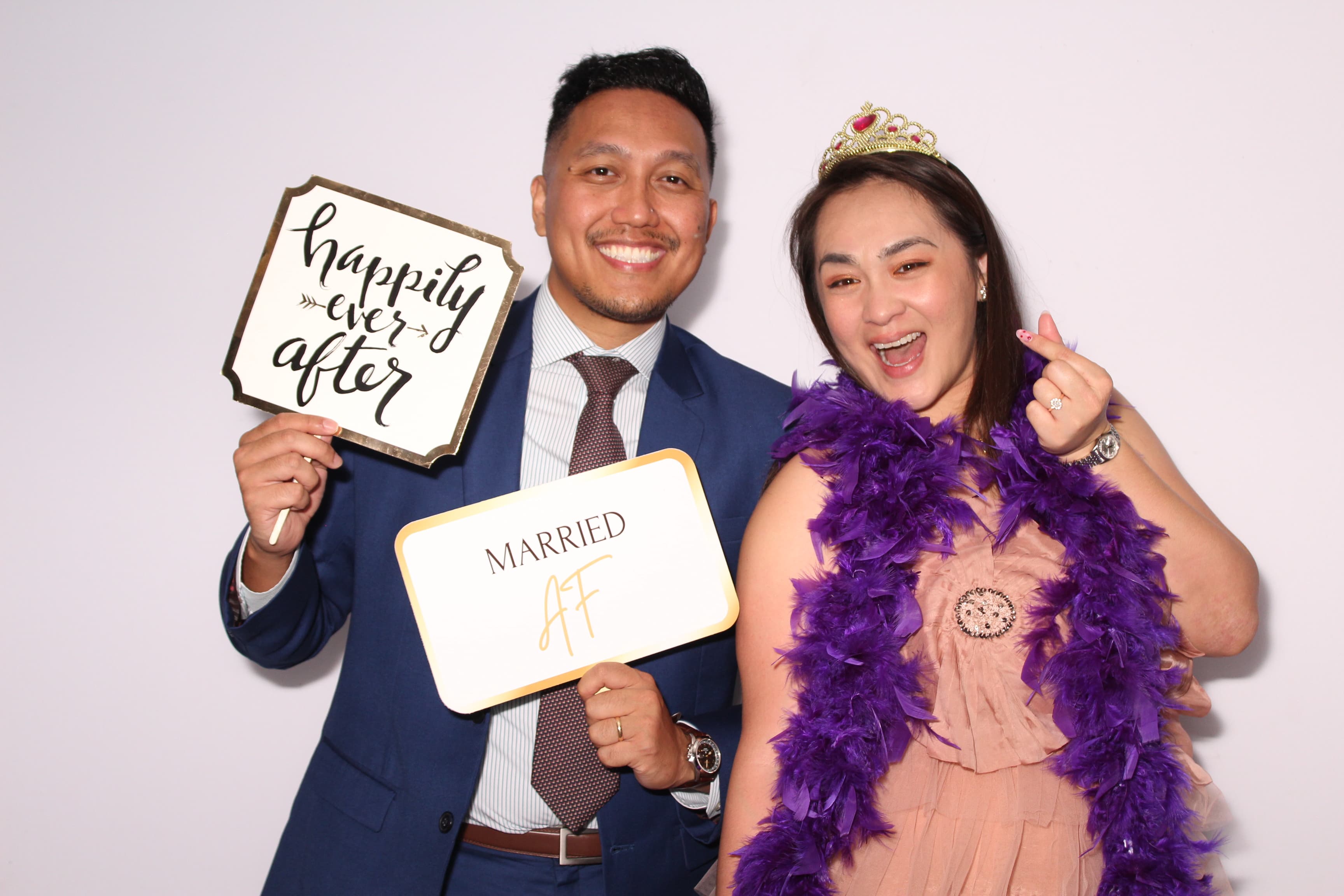 A couple celebrating with ‘Happily Ever After’ and ‘Married AF’ signs, the bride in a tiara and purple feather boa.