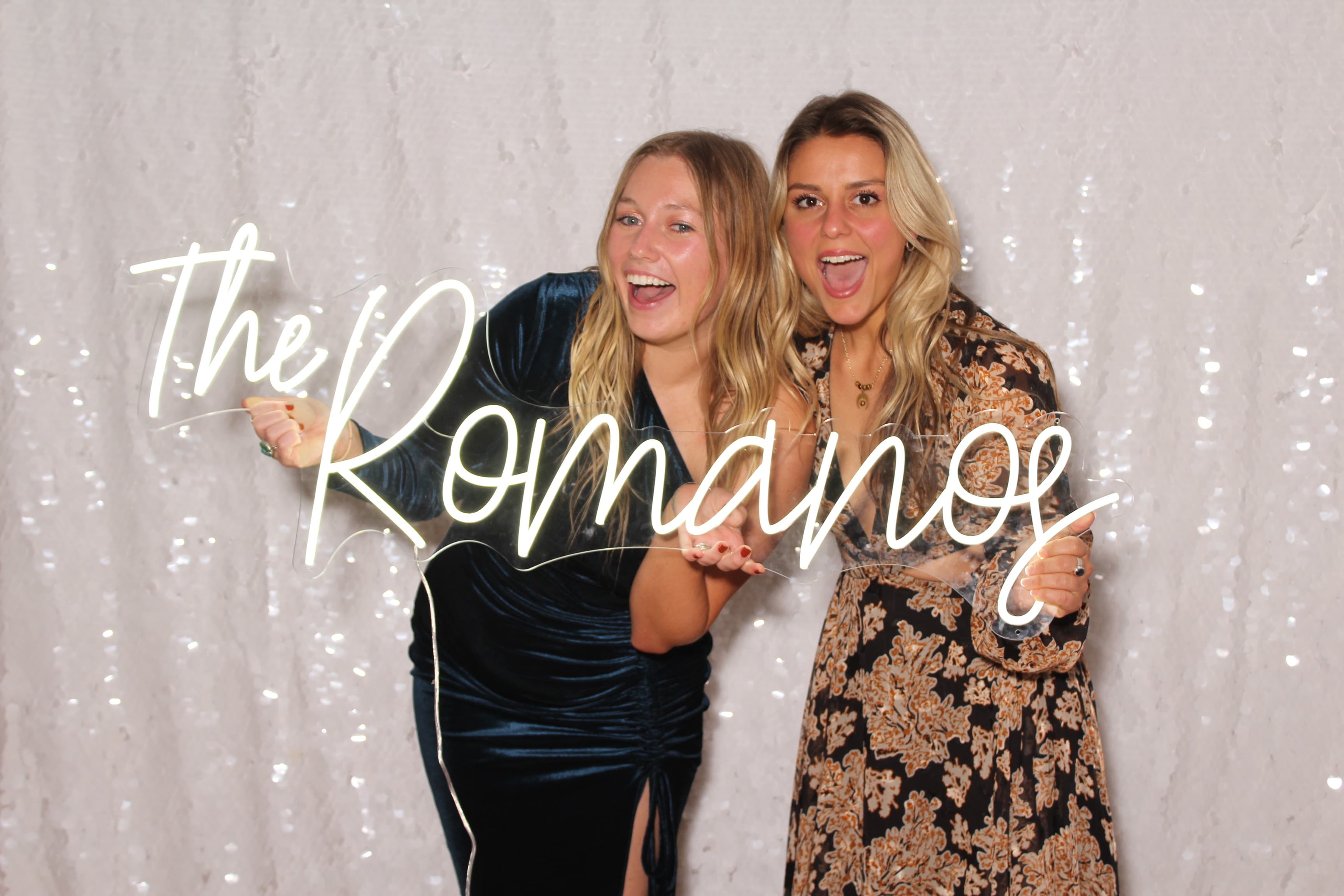 Two friends laughing while holding a glowing neon ‘The Romanos’ sign at a wedding reception.