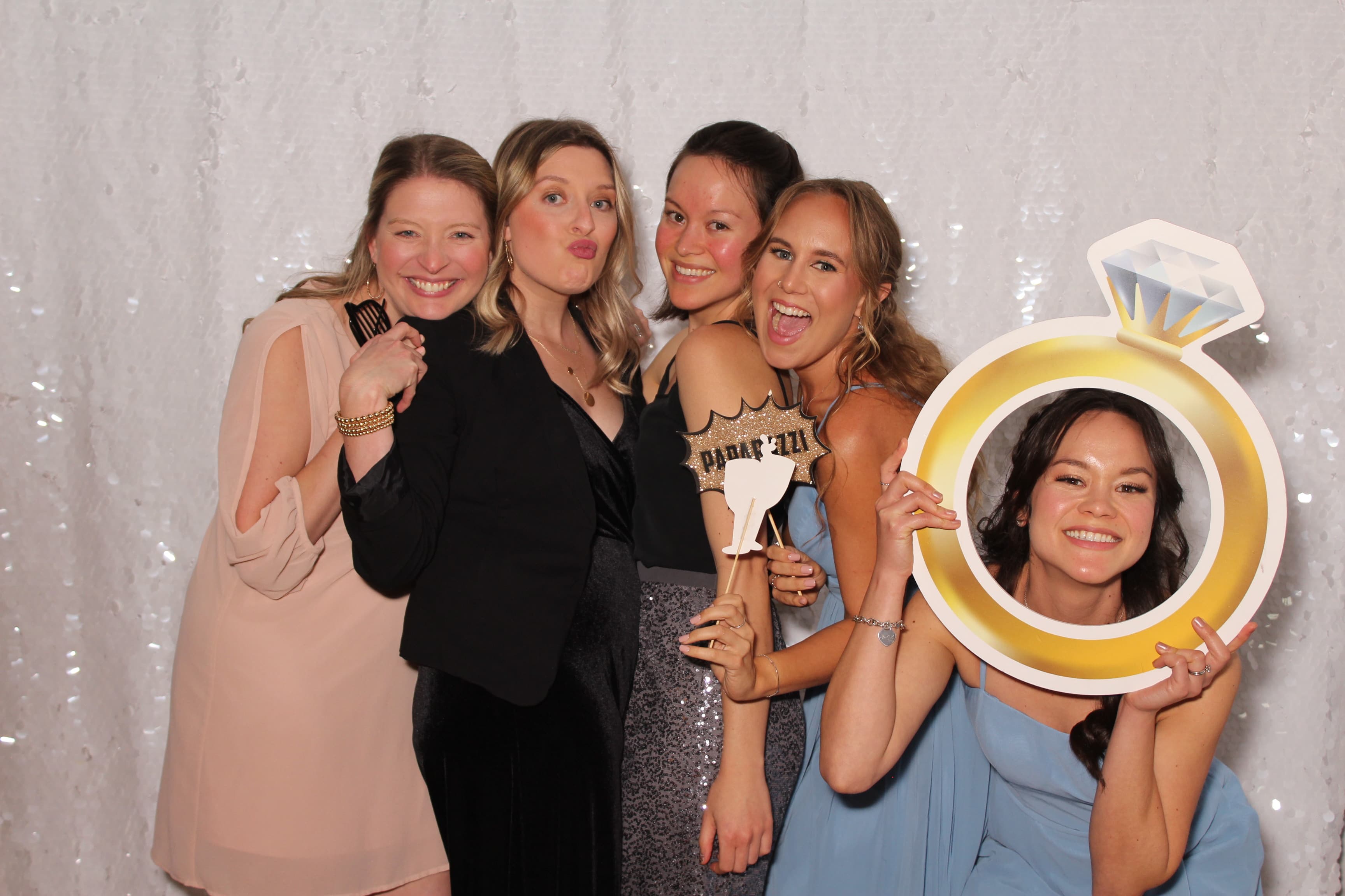 Five friends posing with a paparazzi sign and a giant engagement-ring cutout prop at a bridal celebration.
