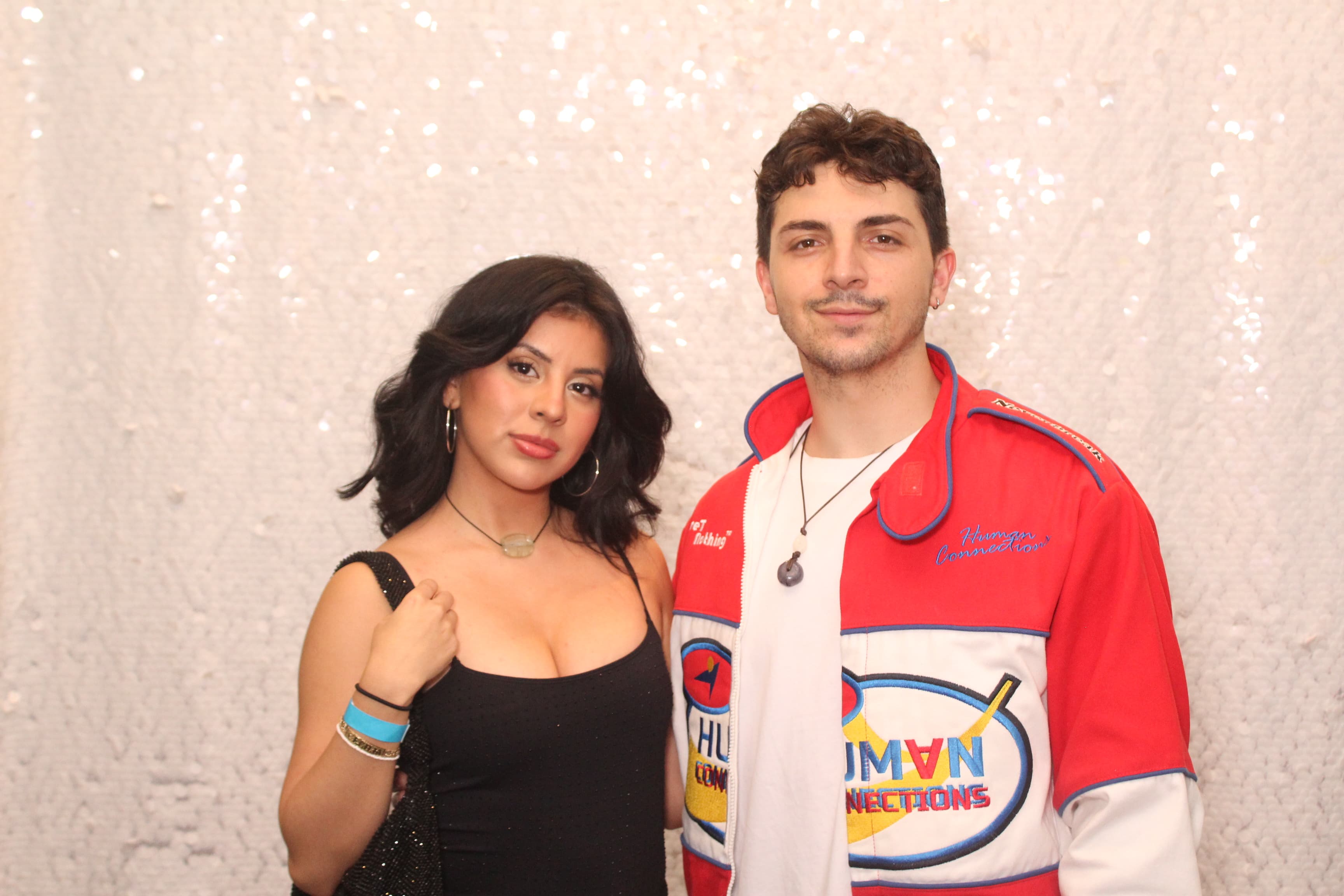 A couple posing together in front of a sparkling sequin backdrop, one wearing a colorful retro racing jacket.