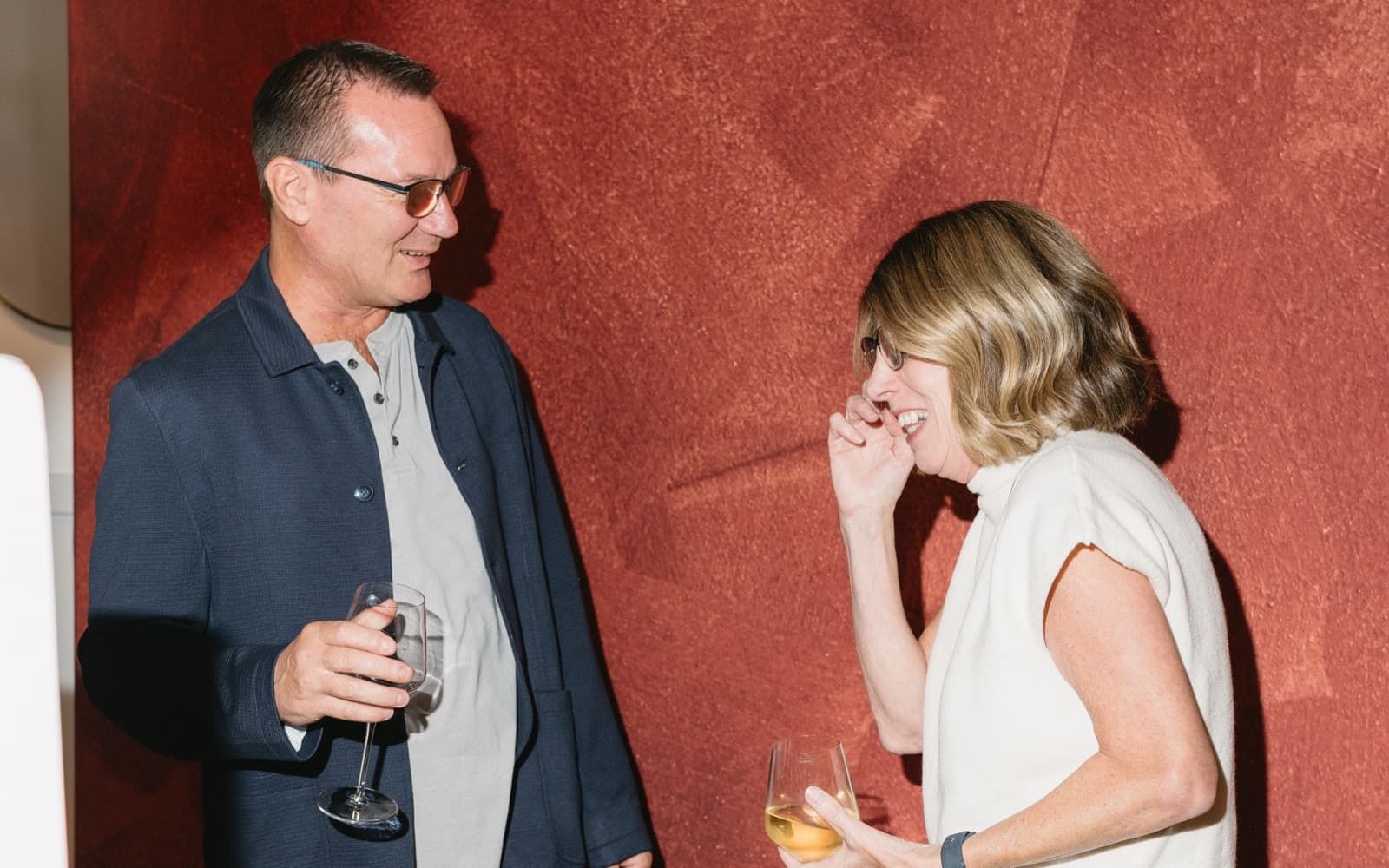 A man and a woman laughing together with wine glasses in front of a rust-red textured backdrop at the MEMORI booth.