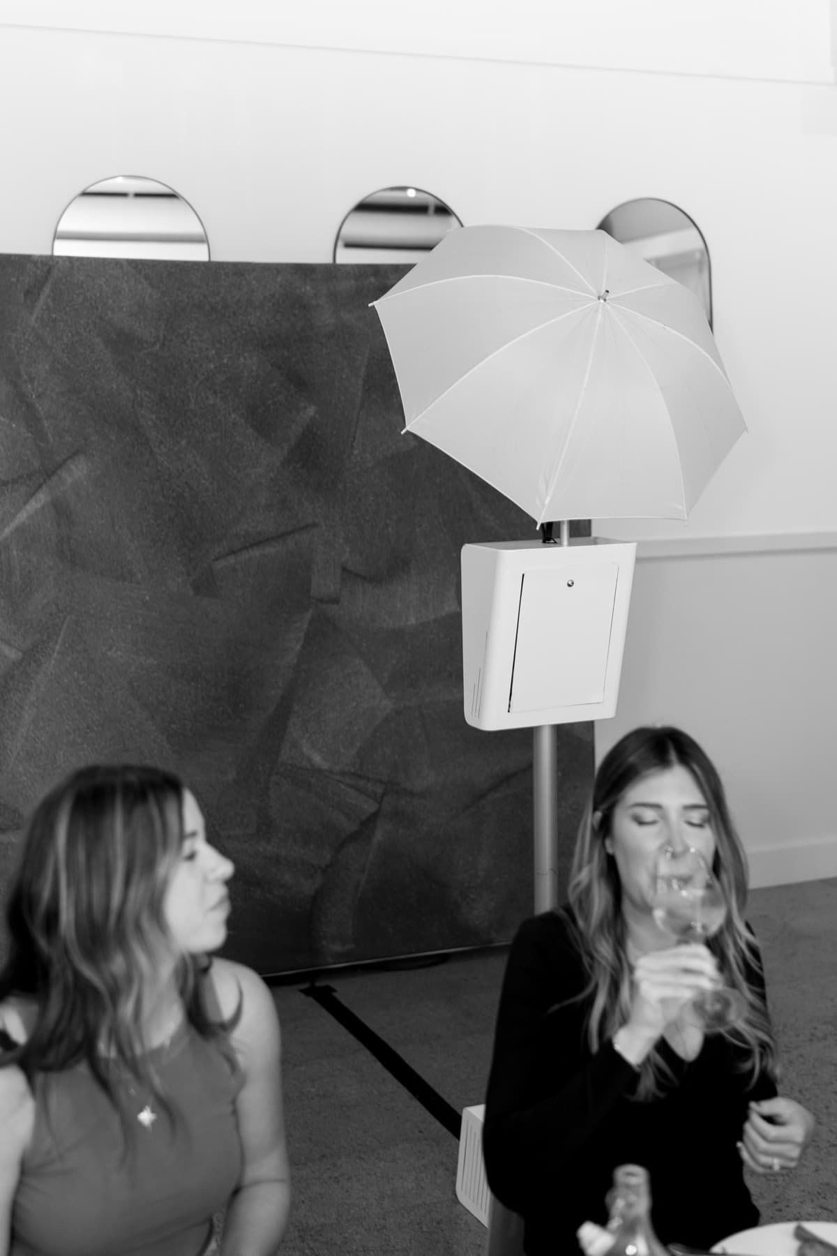 Two friends sipping wine at dinner with the MEMORI photo booth and its soft umbrella light behind them.